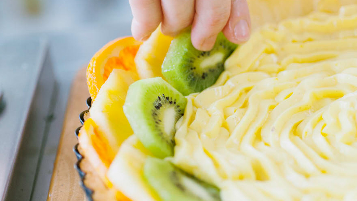 Una mano dispone fette di kiwi sulla crostata già decorata con crema pasticcera a spirale e arancia