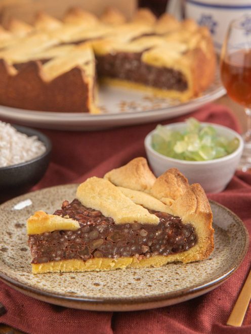 Torta coi bischeri con una fetta servita al piatto, ripieno di cioccolato e pinoli ben visibile