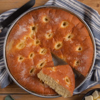 Focaccia dolce dorata con fossette e zucchero semolato, una fetta staccata che mostra la mollica soffice