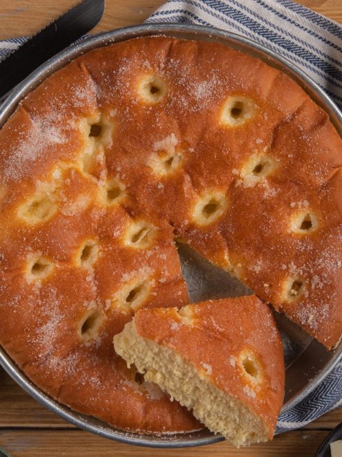 Focaccia dolce dorata con fossette e zucchero semolato, una fetta staccata che mostra la mollica soffice