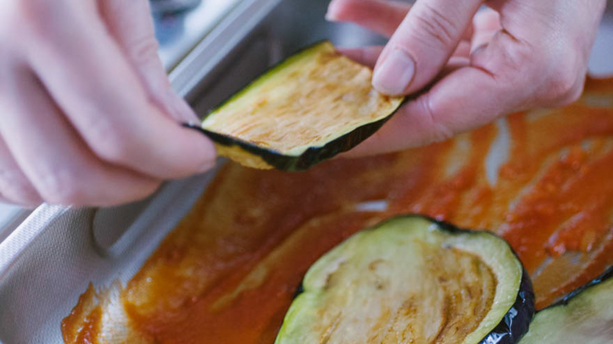 Mani che adagiano una fetta di melanzana fritta sullo strato di sugo nella teglia