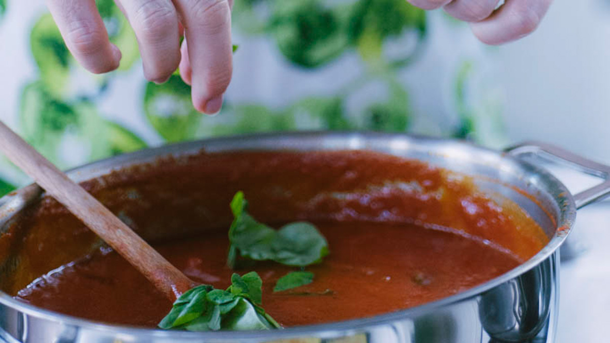 Foglie di basilico fresco aggiunte a mano in un sugo di pomodoro in cottura, mescolato con un cucchiaio di legno