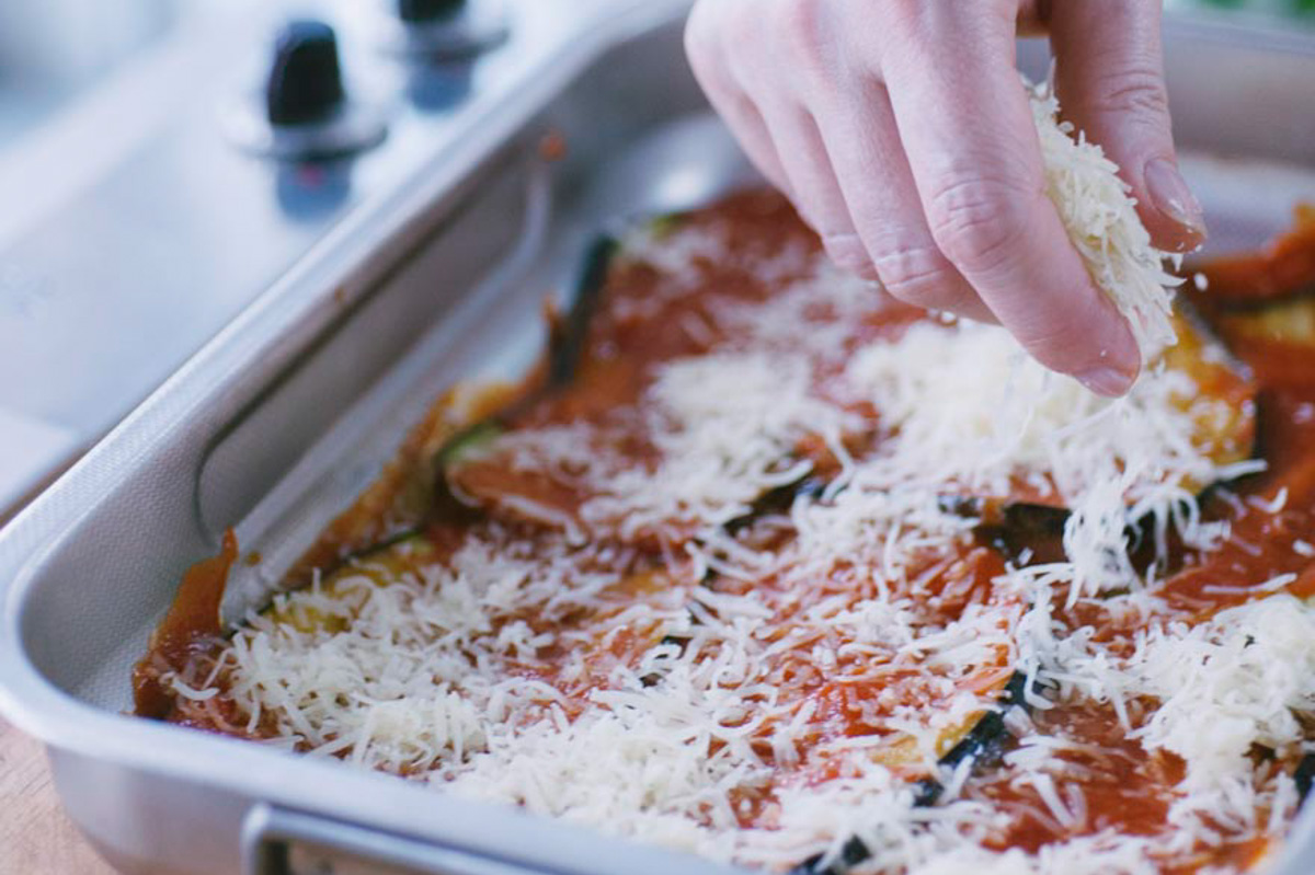 Mano che sparge formaggio grattugiato sulla parmigiana di melanzane a strati con salsa di pomodoro
