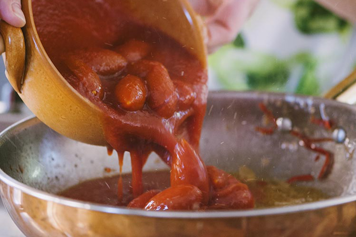 Pomodori pelati versati in padella dove sfrigola il condimento dell'amatriciana