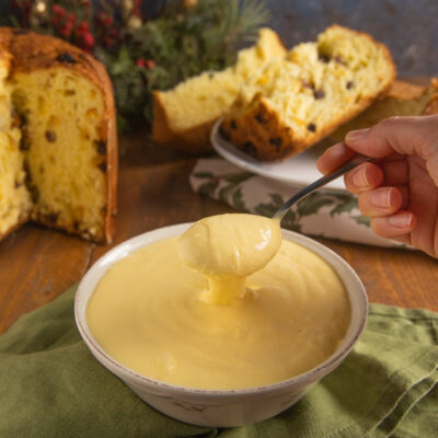 Crema al mascarpone servita in una ciotola con un cucchiaio che la solleva, e fette di panettone sullo sfondo