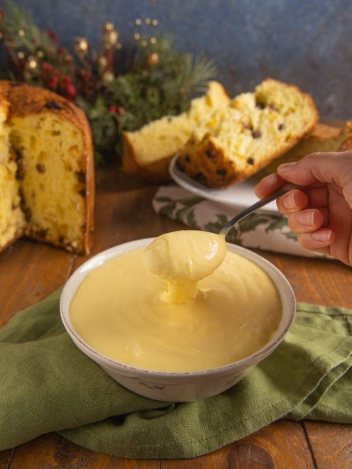 Crema al mascarpone servita in una ciotola con un cucchiaio che la solleva, e fette di panettone sullo sfondo