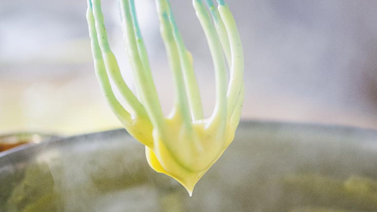 Crema pasticcera densa che cola dalla frusta, con consistenza vellutata e colore giallo paglierino