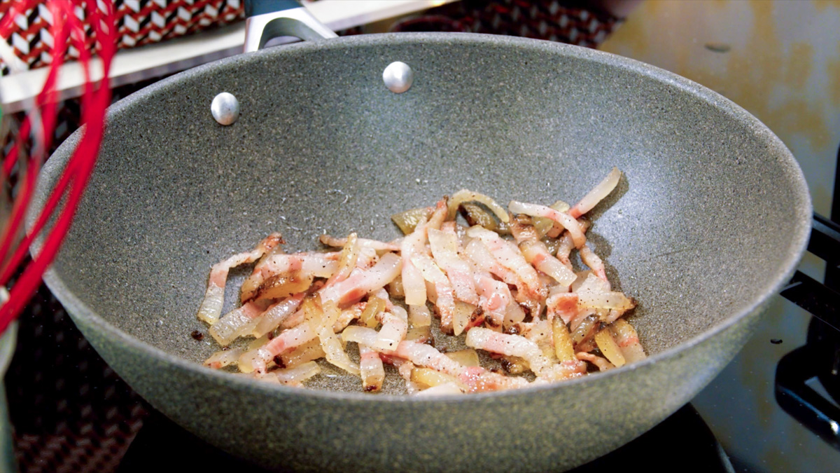 Guanciale a listarelle che rosola con cipolla in padella, pepe nero in grana visibile