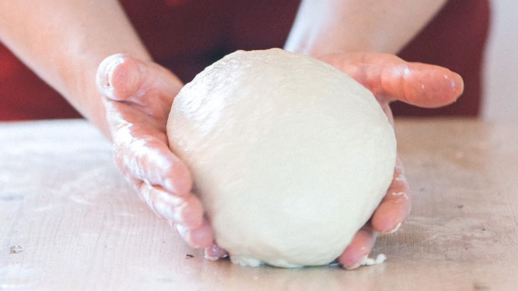 Mani che modellano un panetto di impasto per pizza liscio ed elastico sul piano infarinato