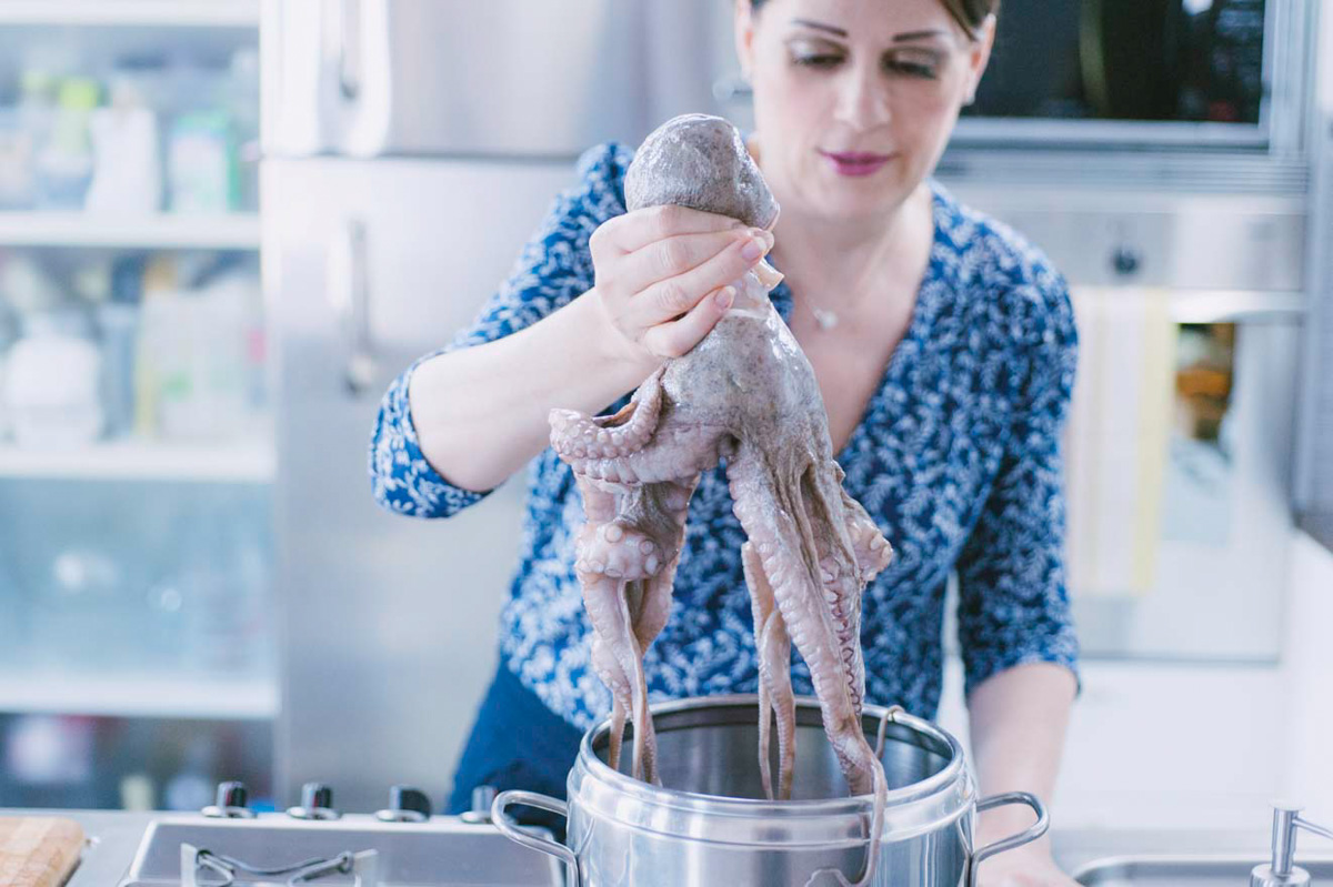 Mano che tiene un polpo crudo per la testa sopra una pentola, pronta a immergere i tentacoli