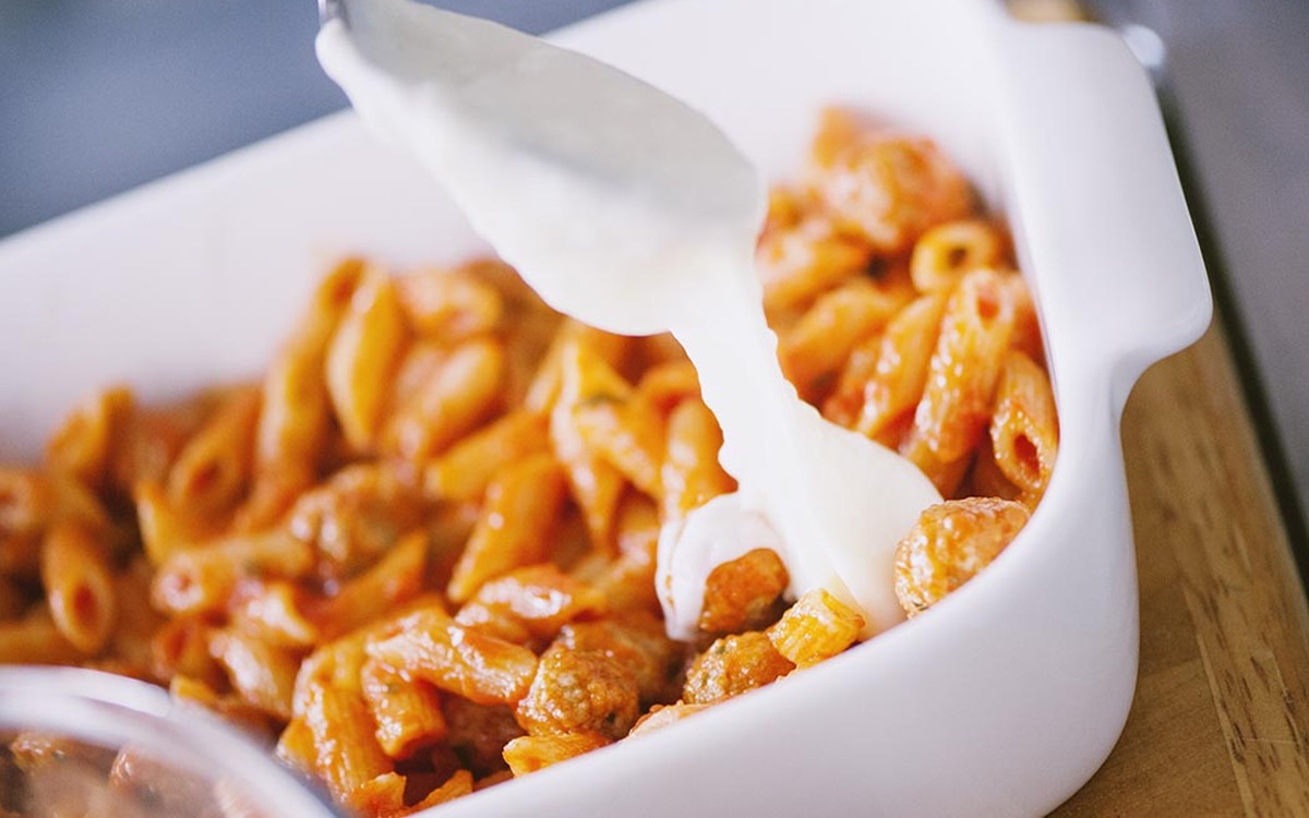 Besciamella versata a filo sulle penne al sugo di carne nella teglia, pronta per la stratificazione