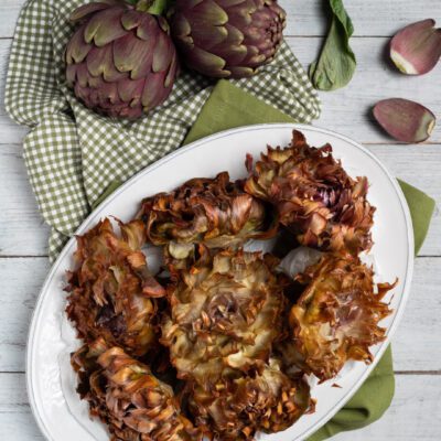 Carciofi alla Giudia fritti e aperti a fiore, serviti su vassoio con carciofi freschi sullo sfondo