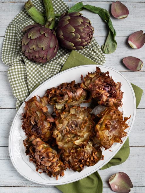 Carciofi alla Giudia fritti e aperti a fiore, serviti su vassoio con carciofi freschi sullo sfondo