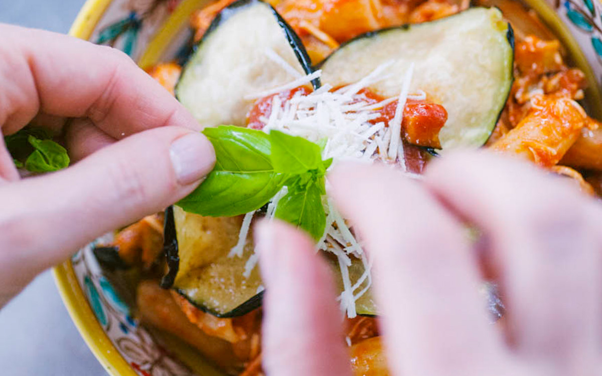 Mani che adagiano foglie di basilico fresco sulla pasta alla norma con melanzane e ricotta salata grattugiata