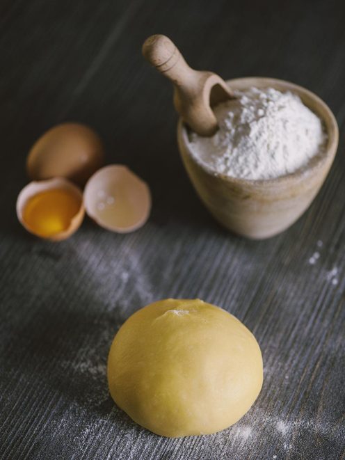 Pasta fresca all'uovo: palla di impasto giallo dorato con farina e uova aperte sullo sfondo