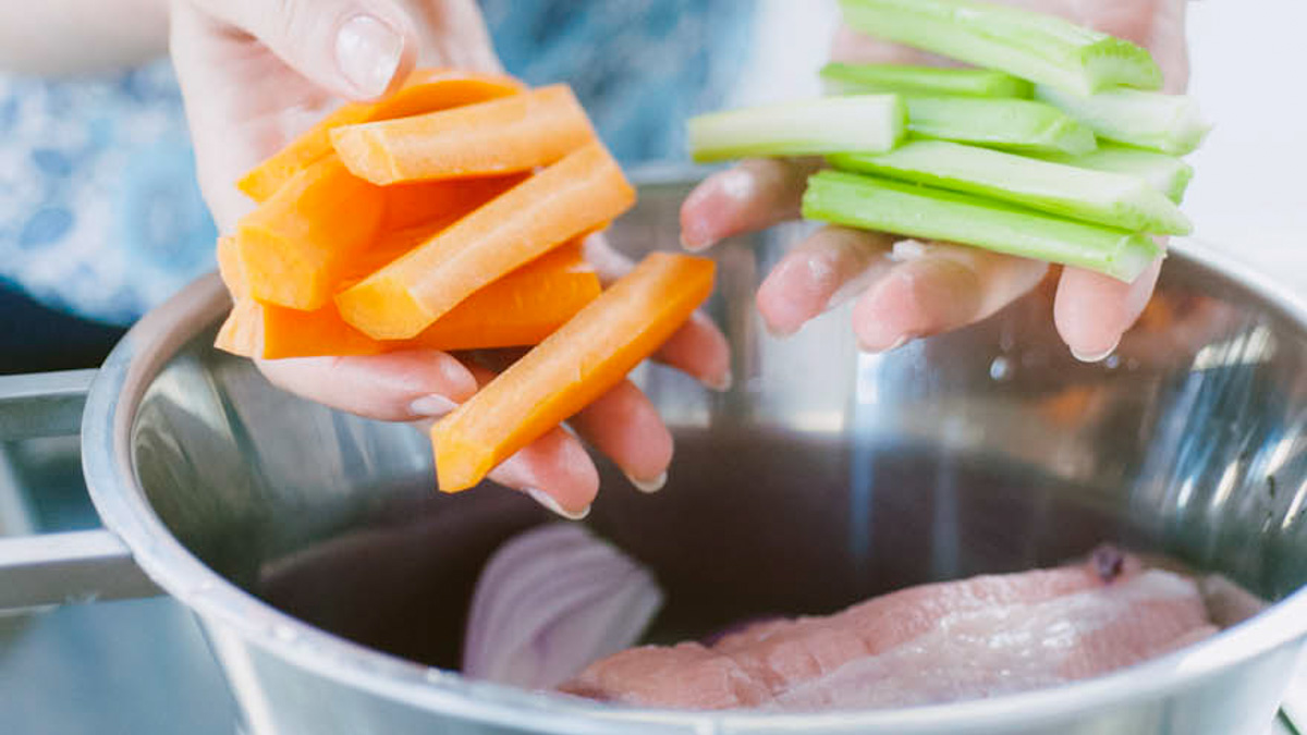 Mani che aggiungono carote e sedano a bastoncini in una pentola con carne e cipolla