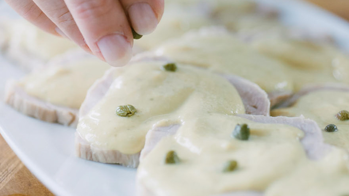 Mano che dispone capperi sulla salsa tonnata che ricopre le fette di vitello
