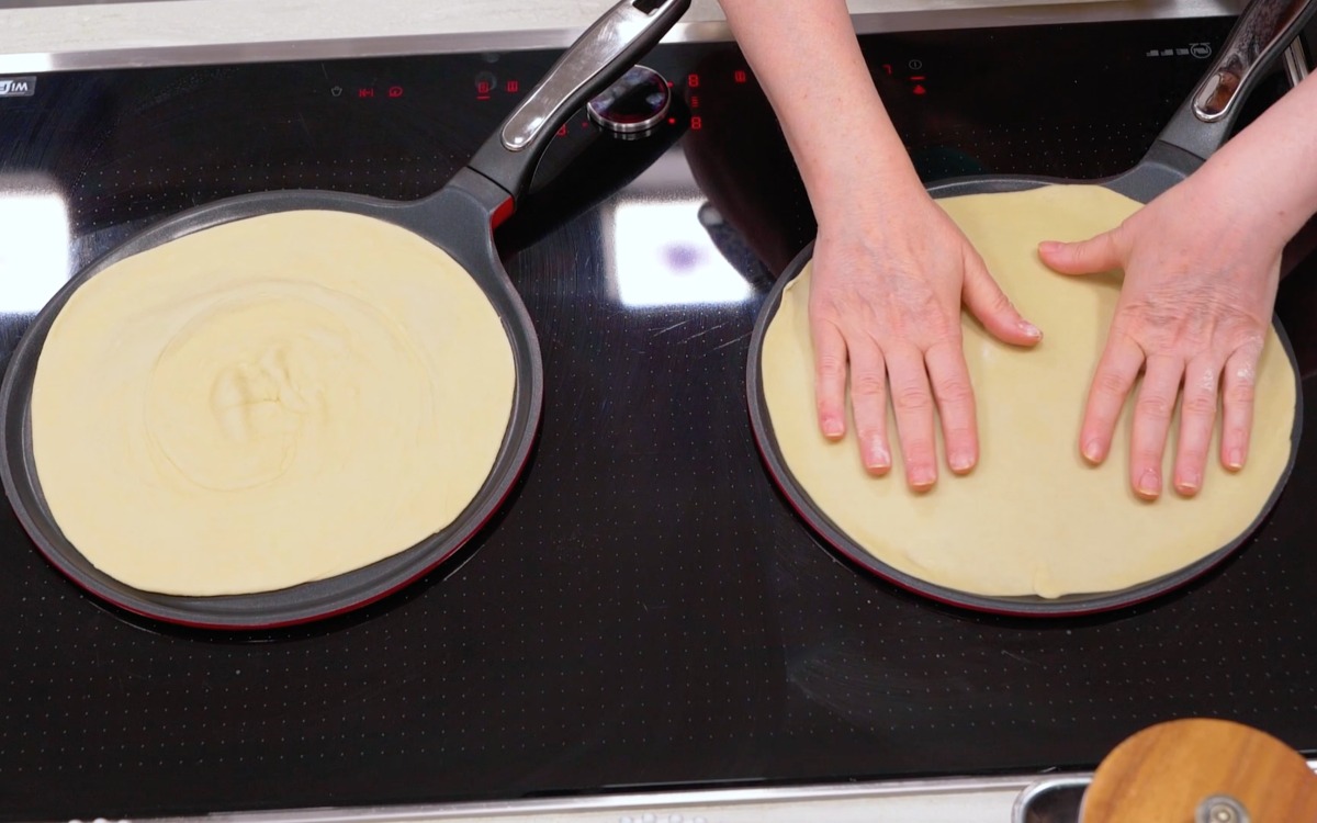 Mani che appiattiscono un disco di pasta cruda su una piastra, mentre un altro cuoce accanto