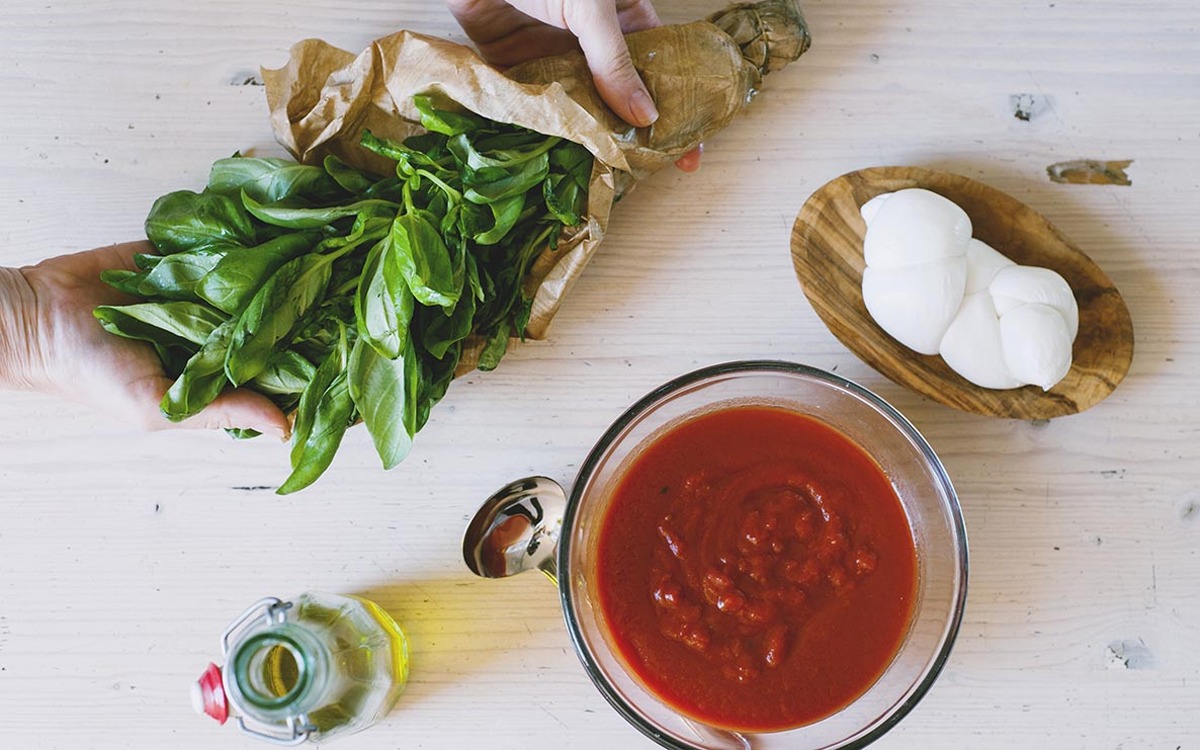 Mano che regge un mazzo di basilico fresco accanto a salsa di pomodoro rustica e mozzarella