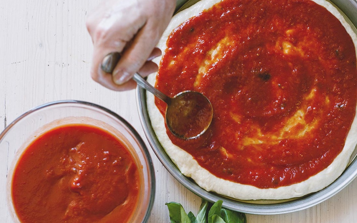 Salsa di pomodoro spalmata con un mestolo sulla base della pizza, bordo bianco lasciato libero