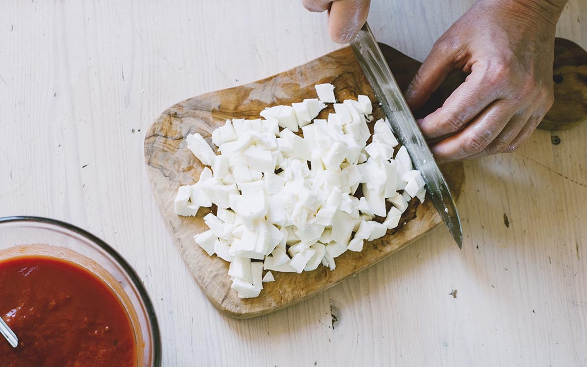 Mani che tagliano a cubetti il fiordilatte su un tagliere, con salsa di pomodoro sullo sfondo