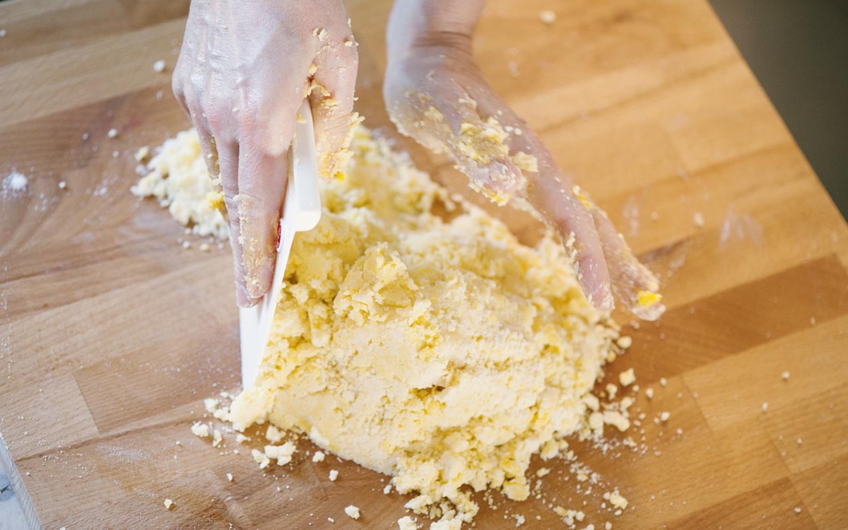 Mani che compattano la pasta frolla sbriciolata sul piano di lavoro con un tarocco