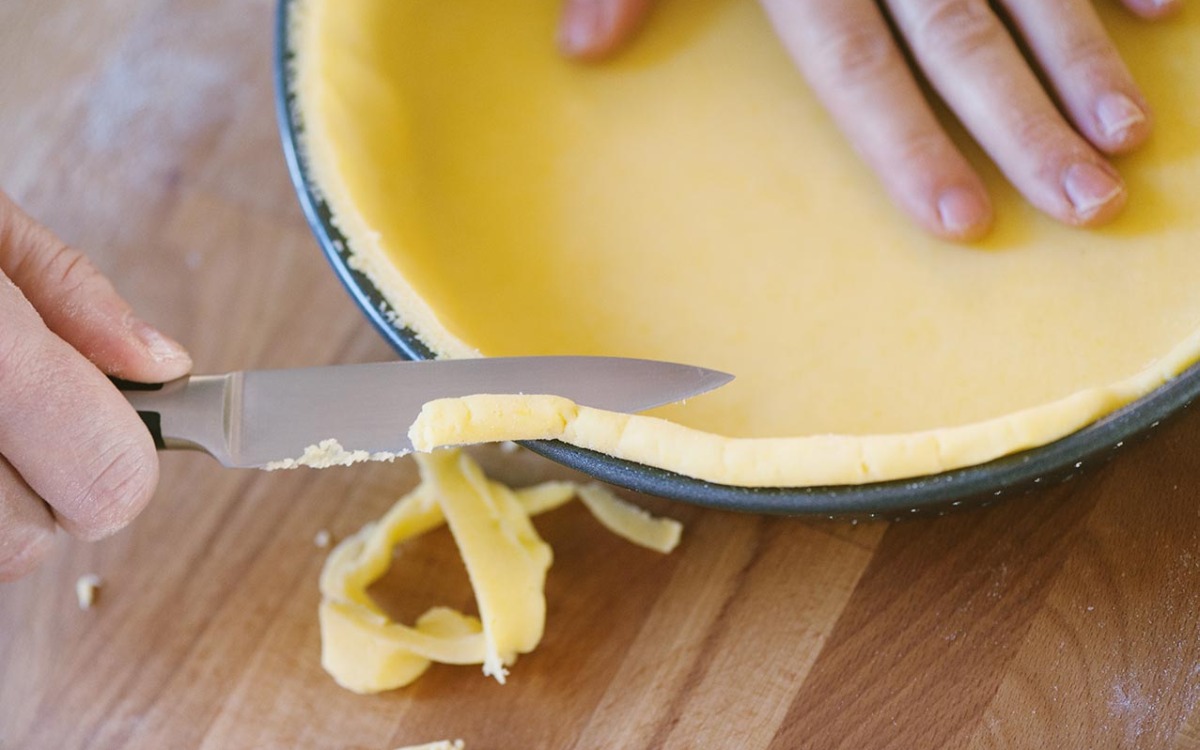 Pasta frolla stesa nella tortiera mentre un coltello taglia via l'eccesso dai bordi