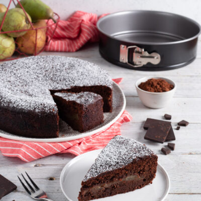 Torta pere e cioccolato spolverata di zucchero a velo, con una fetta servita che rivela il ripieno di pere