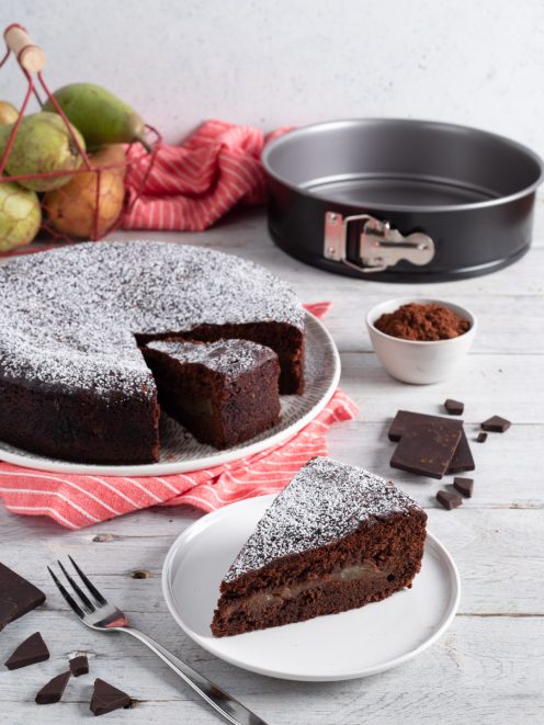 Torta pere e cioccolato spolverata di zucchero a velo, con una fetta servita che rivela il ripieno di pere