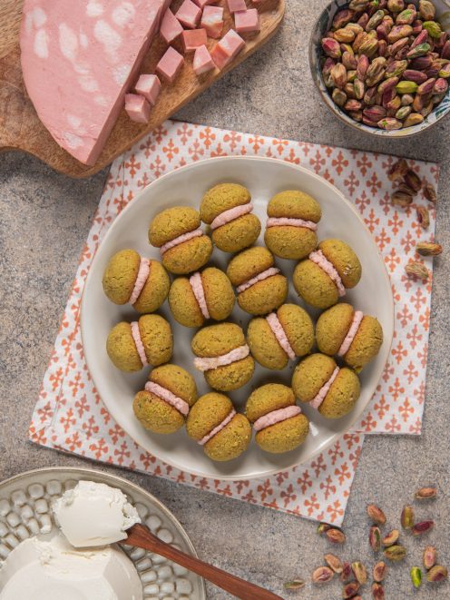 Baci di dama salati con mousse di mortadella disposti su piatto e tovagliolo bianco e rosa, con mortadella a dadini e pistacchi