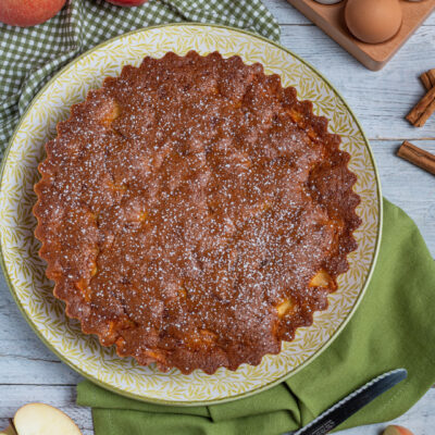 Torta rustica alle mele dorata e spolverata di zucchero a velo, circondata da mele fresche e uova