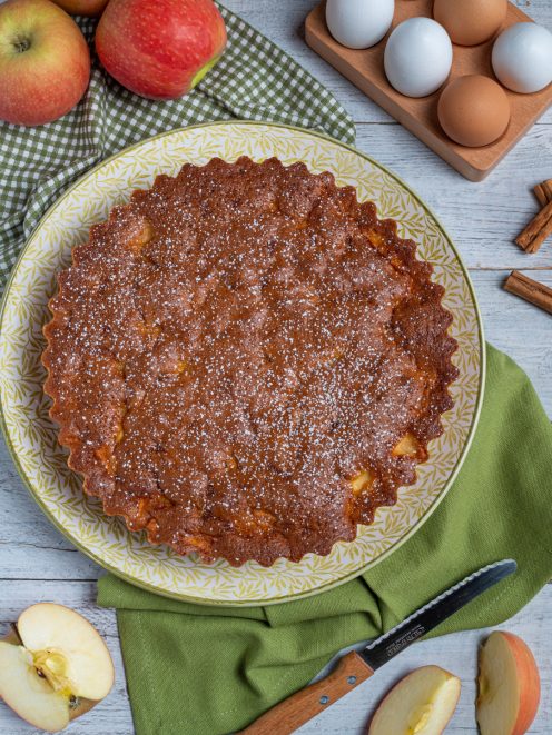 Torta rustica alle mele dorata e spolverata di zucchero a velo, circondata da mele fresche e uova
