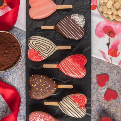 Tortine a cuore su stecco glassate con cioccolato fondente, bianco e rosa, decorate con codette e perline