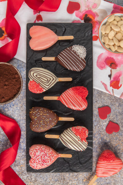 Tortine a cuore su stecco glassate con cioccolato fondente, bianco e rosa, decorate con codette e perline