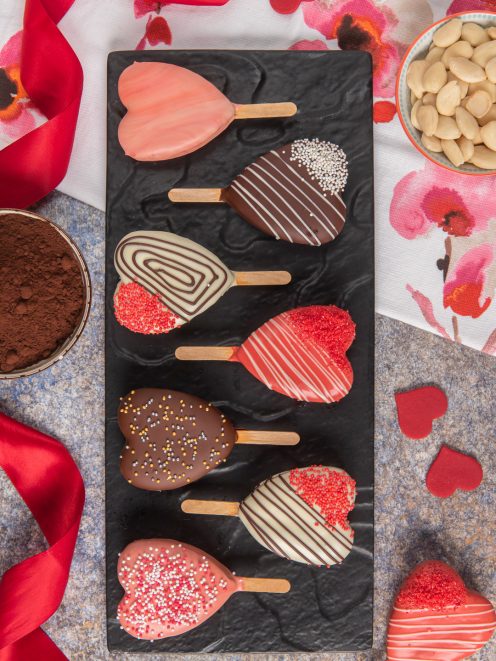 Tortine a cuore su stecco glassate con cioccolato fondente, bianco e rosa, decorate con codette e perline