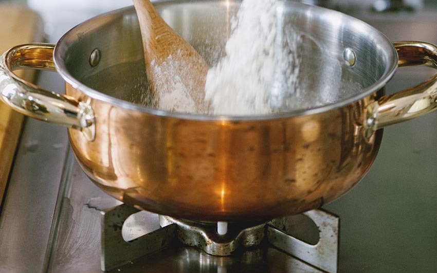 Farina versata in un pentolino sul fuoco mentre un cucchiaio di legno mescola il roux
