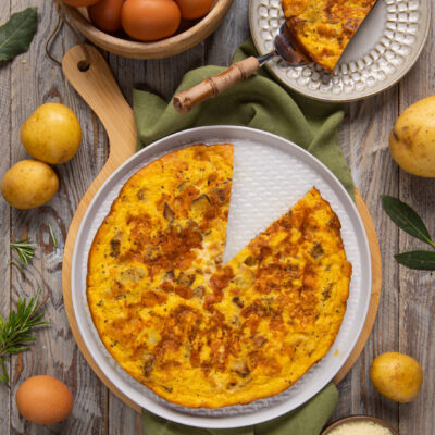 Frittata di patate dorata con una fetta sollevata, circondata da uova, patate e parmigiano grattugiato