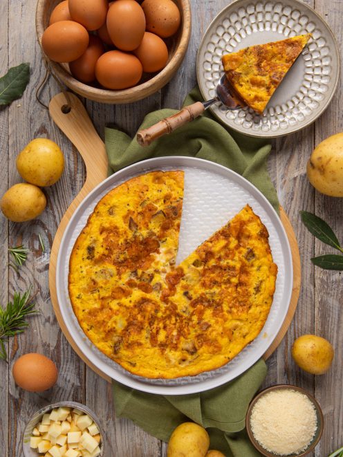 Frittata di patate dorata con una fetta sollevata, circondata da uova, patate e parmigiano grattugiato