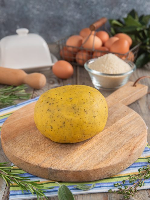 Pasta frolla salata alle erbe aromatiche pronta per essere stesa, con uova e farina sullo sfondo