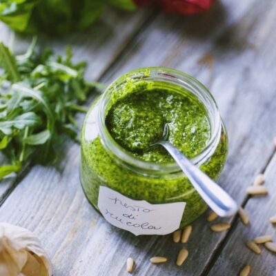 Pesto di rucola in barattolo con cucchiaino, circondato da rucola fresca, aglio e pinoli