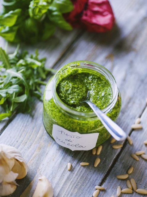 Pesto di rucola in barattolo con cucchiaino, circondato da rucola fresca, aglio e pinoli
