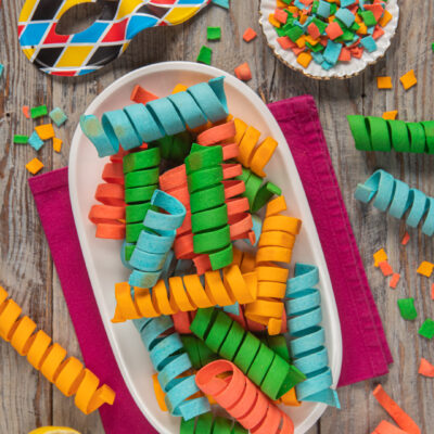 Stelle filanti di pasta colorata a spirale su piatto ovale, tra coriandoli e maschera di carnevale