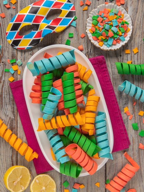 Stelle filanti di pasta colorata a spirale su piatto ovale, tra coriandoli e maschera di carnevale