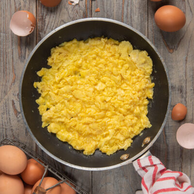 Uova strapazzate morbide e cremose in padella, circondate da uova fresche e gusci rotti