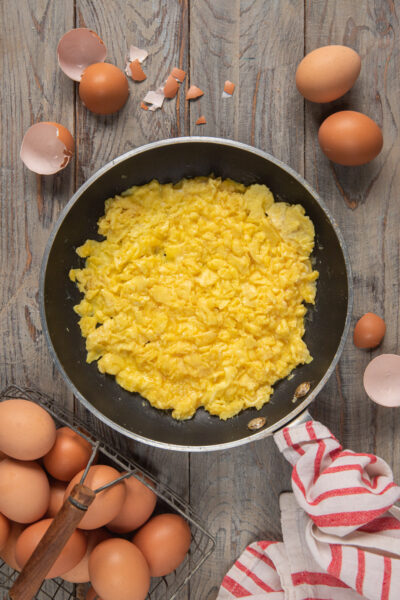 Uova strapazzate morbide e cremose in padella, circondate da uova fresche e gusci rotti