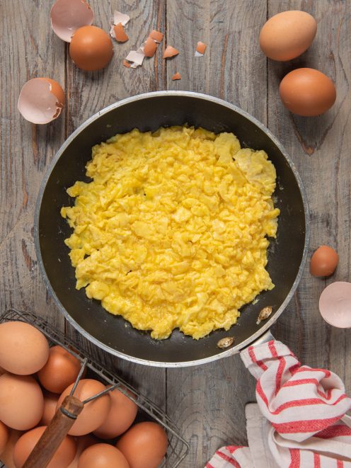 Uova strapazzate morbide e cremose in padella, circondate da uova fresche e gusci rotti