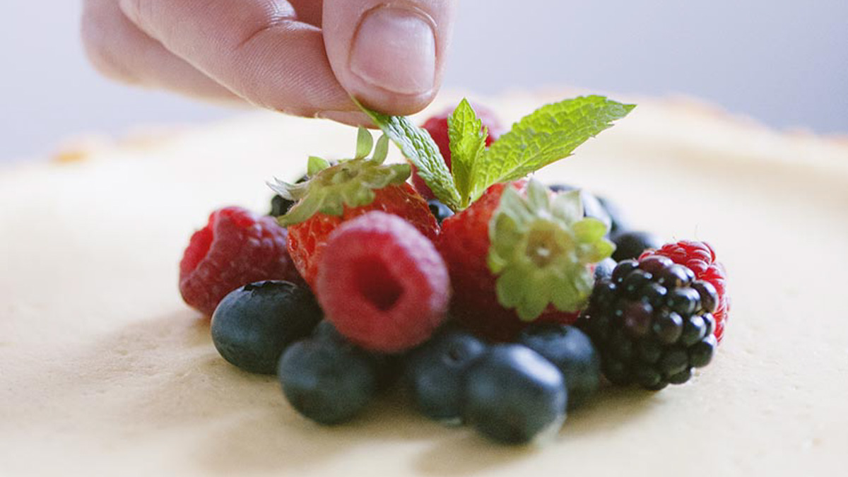 Mano che adagia foglioline di menta su frutti di bosco freschi sopra la cheesecake