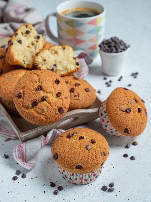 Muffin con gocce di cioccolato disposti su vassoio con uno tagliato a meta e tazza di caffe