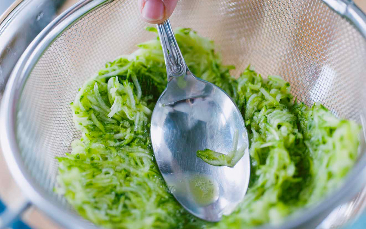 Zucchine grattugiate scolate in un colino mentre una mano le preme con un cucchiaio