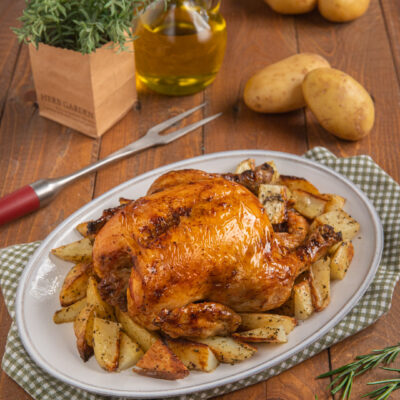 Pollo arrosto intero con pelle dorata e croccante, circondato da patate al forno a spicchi
